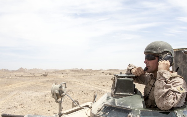A military service member in desert camouflage uniform and helmet is positioned in the open hatch of an armored vehicle, using a radio handset to communicate. The scene is set in an arid desert environment with flat terrain and distant rocky formations visible in the background. The armored vehicle shows signs of heavy use and wear.