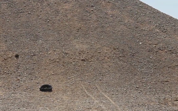 The image shows an arid desert landscape with a large rocky hill or mountain slope in the background. Several dark objects are scattered across the flat desert floor, with visible dust disturbance suggesting recent activity. The scene appears to capture some form of impact or dispersal event in a remote desert environment.