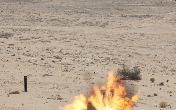 An explosion is occurring on a flat, arid desert terrain with sparse scrub vegetation. The detonation appears to be a controlled demolition or ordnance disposal event, evidenced by a marker post visible to the left of the blast. The image was captured at the moment of detonation, showing active flames and debris ejection.