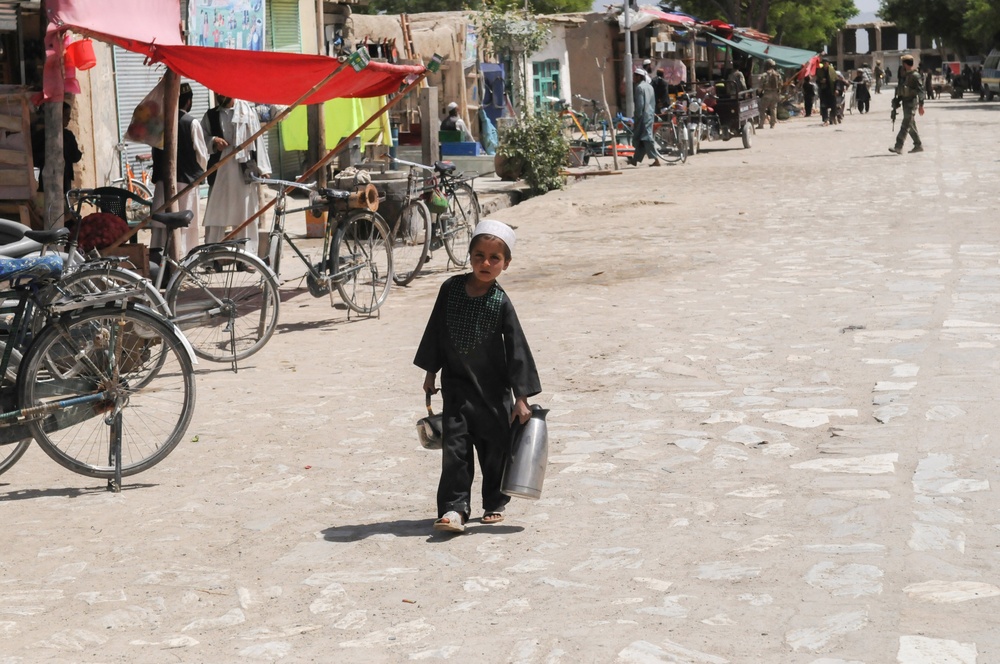 A young child dressed in traditional dark shalwar kameez and white taqiyah (cap) walks along a dusty market street carrying two metal canisters or containers. The scene depicts a busy bazaar environment with bicycles, rickshaws, market stalls with colorful awnings, and various people in the background. The setting appears to be a rural or semi-urban marketplace in South or Central Asia, likely Afghanistan or Pakistan.