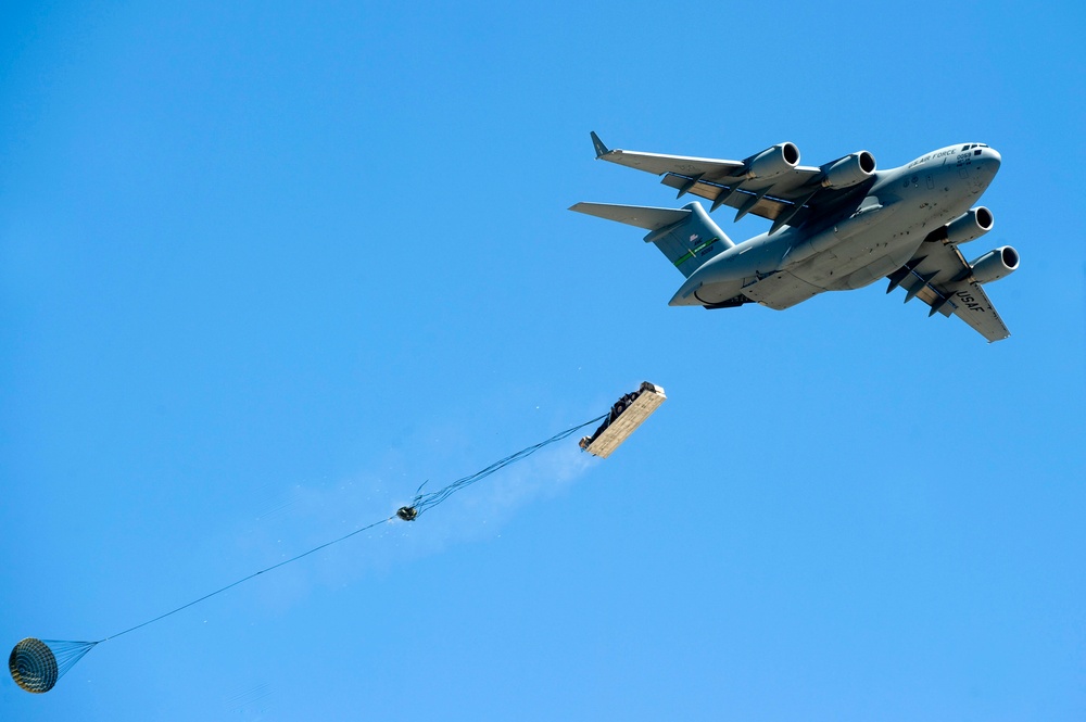 A U.S. Air Force C-17 Globemaster III military transport aircraft is performing an aerial cargo airdrop operation against a clear blue sky. A large pallet of cargo has been released from the aircraft's rear and is being slowed by a deployed extraction/drogue parachute connected via a long tether. The cargo appears to be emitting smoke or dust upon release, consistent with a heavy equipment airdrop.