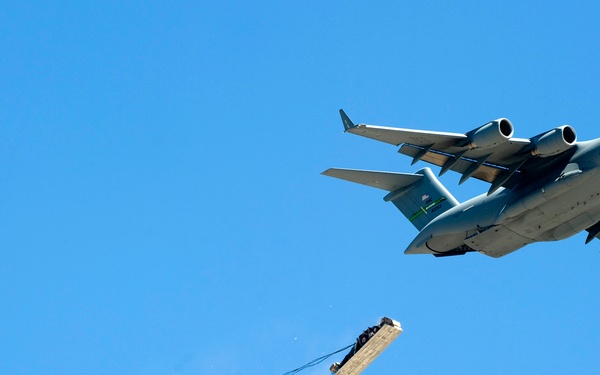 A U.S. Air Force C-17 Globemaster III military transport aircraft is performing an aerial cargo airdrop operation against a clear blue sky. A large pallet of cargo has been released from the aircraft's rear and is being slowed by a deployed extraction/drogue parachute connected via a long tether. The cargo appears to be emitting smoke or dust upon release, consistent with a heavy equipment airdrop.