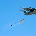 A U.S. Air Force C-17 Globemaster III military transport aircraft is performing an aerial cargo airdrop operation against a clear blue sky. A large pallet of cargo has been released from the aircraft's rear and is being slowed by a deployed extraction/drogue parachute connected via a long tether. The cargo appears to be emitting smoke or dust upon release, consistent with a heavy equipment airdrop.