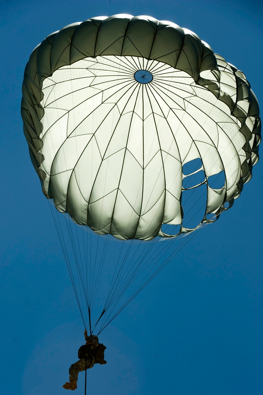 A military paratrooper is descending under a fully deployed round parachute against a clear blue sky. The parachute appears to be an olive drab and white military-style canopy with visible vents and suspension lines. The image is taken from below, looking upward at the parachutist in silhouette.