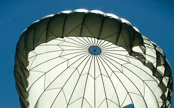A military paratrooper is descending under a fully deployed round parachute against a clear blue sky. The parachute appears to be an olive drab and white military-style canopy with visible vents and suspension lines. The image is taken from below, looking upward at the parachutist in silhouette.