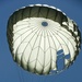A military paratrooper is descending under a fully deployed round parachute against a clear blue sky. The parachute appears to be an olive drab and white military-style canopy with visible vents and suspension lines. The image is taken from below, looking upward at the parachutist in silhouette.