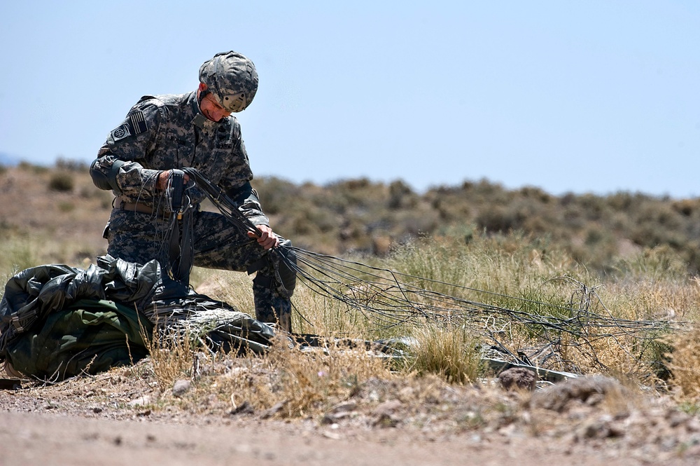 A U.S. Army soldier in ACU (Army Combat Uniform) is gathering and recovering parachute suspension lines after a landing in an arid, desert-scrub terrain. The soldier is kneeling and bundling the lines by hand, with the collapsed parachute canopy visible on the ground nearby. The setting appears to be a training or operational drop zone in a semi-arid environment.