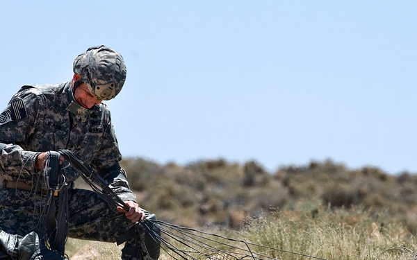 A U.S. Army soldier in ACU (Army Combat Uniform) is gathering and recovering parachute suspension lines after a landing in an arid, desert-scrub terrain. The soldier is kneeling and bundling the lines by hand, with the collapsed parachute canopy visible on the ground nearby. The setting appears to be a training or operational drop zone in a semi-arid environment.