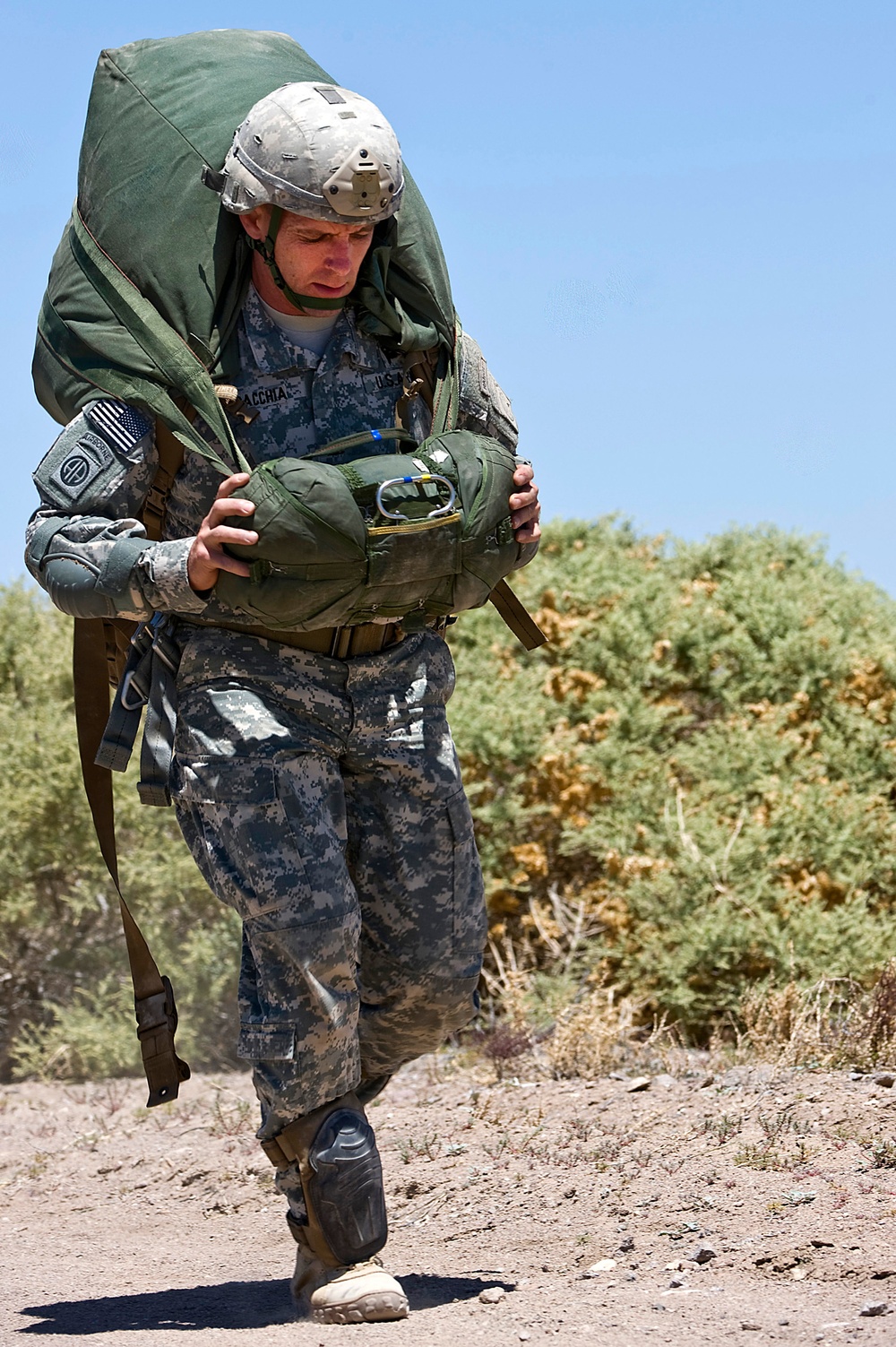 A U.S. Army soldier in Army Combat Uniform (ACU) digital camouflage is carrying a large, heavily loaded parachute pack and rucksack across an arid, desert-like terrain. The soldier is wearing a MICH/ACH combat helmet and bears a visible 82nd Airborne Division patch on the left shoulder. The image appears to capture a post-jump recovery or ruck march exercise under bright, sunny conditions.