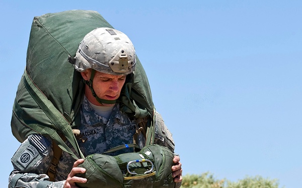 A U.S. Army soldier in Army Combat Uniform (ACU) digital camouflage is carrying a large, heavily loaded parachute pack and rucksack across an arid, desert-like terrain. The soldier is wearing a MICH/ACH combat helmet and bears a visible 82nd Airborne Division patch on the left shoulder. The image appears to capture a post-jump recovery or ruck march exercise under bright, sunny conditions.