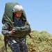 A U.S. Army soldier in Army Combat Uniform (ACU) digital camouflage is carrying a large, heavily loaded parachute pack and rucksack across an arid, desert-like terrain. The soldier is wearing a MICH/ACH combat helmet and bears a visible 82nd Airborne Division patch on the left shoulder. The image appears to capture a post-jump recovery or ruck march exercise under bright, sunny conditions.