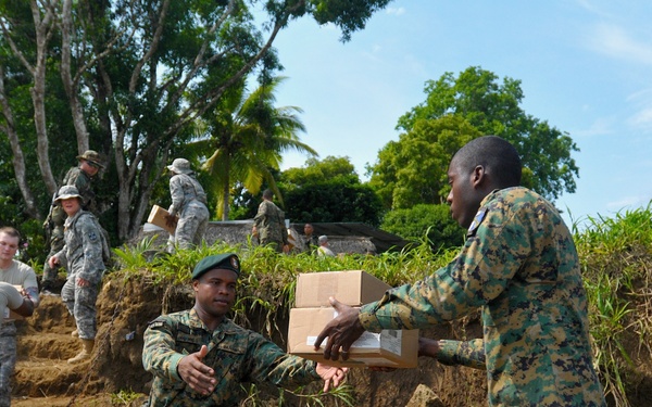 Military personnel from what appear to be two different forces are conducting a supply distribution operation near a riverbank or waterway. Soldiers are forming a human chain to pass cardboard boxes from an elevated embankment down into a wooden boat. The scene suggests a humanitarian aid or logistics resupply mission in a tropical environment.
