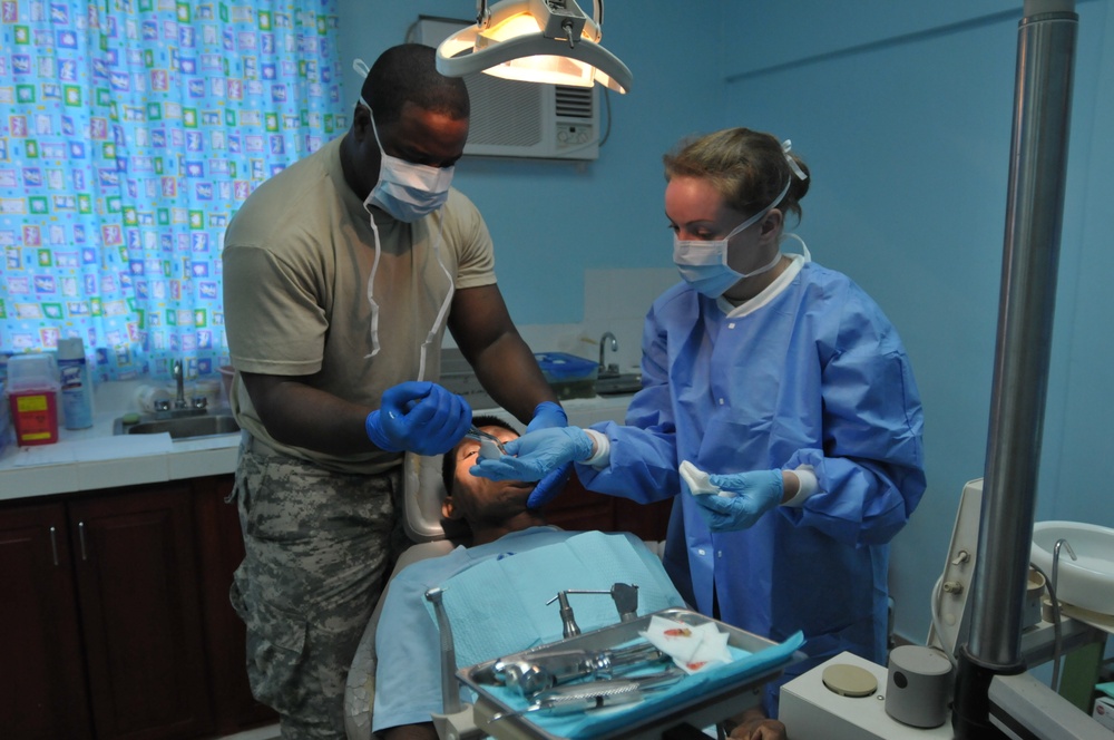 Two medical/dental professionals are performing a dental procedure on a patient reclined in a dental chair. One provider is wearing military camouflage pants and a tan shirt with blue latex gloves, while the other is wearing a full blue surgical gown with gloves and protective eyewear. The setting appears to be a basic dental clinic with blue walls, possibly in a military or humanitarian mission context.