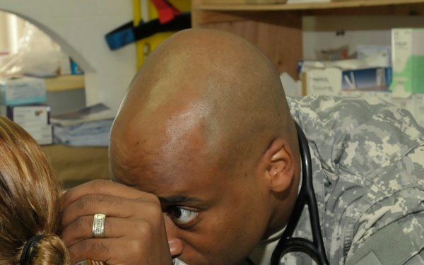 A U.S. Army medical personnel wearing ACU (Army Combat Uniform) is conducting a medical examination on a patient, using an otoscope to examine the patient's ear. The soldier has a stethoscope around his neck and wears a unit patch on his left shoulder. The setting appears to be a military medical clinic or aid station with medical supplies visible in the background.