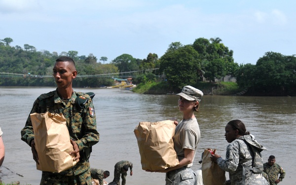 Military personnel from what appear to be two different armed forces are conducting a humanitarian aid or supply distribution operation along a riverbank. Soldiers are carrying brown paper bags and cardboard boxes, forming what appears to be a supply chain from the water's edge. The setting appears to be a tropical or subtropical region based on the lush vegetation and muddy river visible in the background.