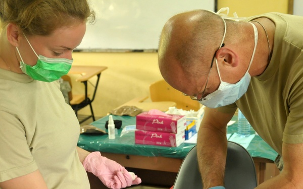 Two military personnel in U.S. Army/Air Force uniforms are providing dental or medical care to a young child seated in a portable dental/medical chair. The setting appears to be a makeshift clinic established in a classroom or community building, consistent with a humanitarian assistance or medical readiness training exercise (MEDRETE) mission. Both providers are wearing surgical masks and latex gloves, indicating a clinical procedure is underway.