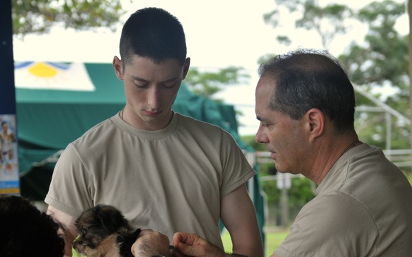 Two individuals in military-style tan/khaki uniforms are examining a small puppy, while a civilian woman in a red and white striped top looks on. The setting appears to be an outdoor community event or veterinary outreach mission, likely in a Latin American country based on the Spanish-language poster visible in the background. The interaction suggests a military humanitarian or veterinary assistance operation.