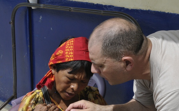 A medical professional is administering an oral medication or vaccine to a small infant using a syringe, while an older indigenous woman holds the baby. The woman is wearing traditional colorful clothing, beaded bracelets, and a red patterned headband, suggesting an indigenous community health outreach setting. The scene appears to take place outdoors or in a semi-outdoor facility with a blue wall in the background.