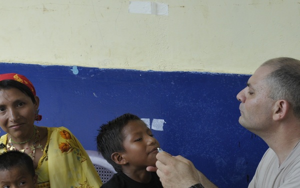 A medical professional appears to be examining or administering oral treatment to a young boy in what looks like a community health setting. A woman, likely the mother, sits nearby holding a toddler dressed in an orange basketball shirt. The setting appears to be a basic facility with a blue and yellow painted wall.
