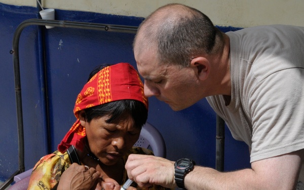 A medical professional is administering an oral medication or vaccine to a young infant being held by a woman wearing traditional indigenous clothing and colorful beaded bracelets. The man is using a small oral syringe to deliver the liquid into the child's mouth. A cardboard box labeled 'BD Syringe' is visible nearby, suggesting a medical outreach or vaccination campaign setting.