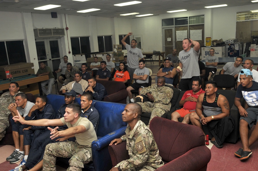 A large group of military personnel and civilians are gathered in what appears to be a military recreation or common room, watching an event off-camera. Several individuals are seated on couches and chairs while two men in Army PT (physical training) shirts are standing and reacting expressively, suggesting they are watching a sporting event. The setting appears to be a military base recreation facility with fluorescent lighting and casual seating arrangements.