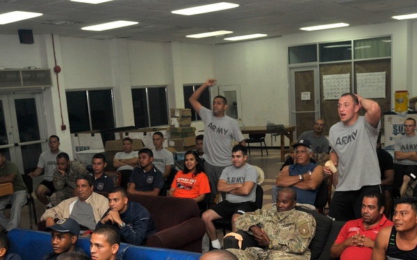 A large group of military personnel and civilians are gathered in what appears to be a military recreation or common room, watching an event off-camera. Several individuals are seated on couches and chairs while two men in Army PT (physical training) shirts are standing and reacting expressively, suggesting they are watching a sporting event. The setting appears to be a military base recreation facility with fluorescent lighting and casual seating arrangements.