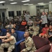 A large group of military personnel and civilians are gathered in what appears to be a military recreation or common room, watching an event off-camera. Several individuals are seated on couches and chairs while two men in Army PT (physical training) shirts are standing and reacting expressively, suggesting they are watching a sporting event. The setting appears to be a military base recreation facility with fluorescent lighting and casual seating arrangements.