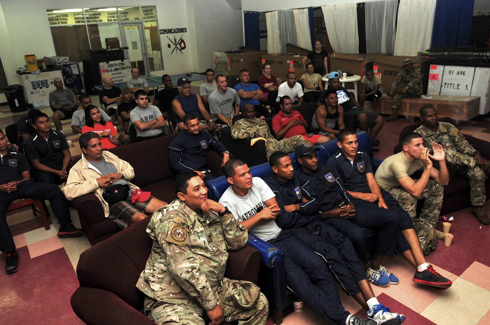 A large group of military personnel and civilians are gathered in what appears to be a common room or recreation area of a military facility. The group is seated on sofas and chairs, appearing to watch something off-camera, possibly a presentation or screen. The setting includes visible military signage in Spanish, including the word 'COMUNICACIONES,' suggesting a Latin American or Spanish-speaking military installation.