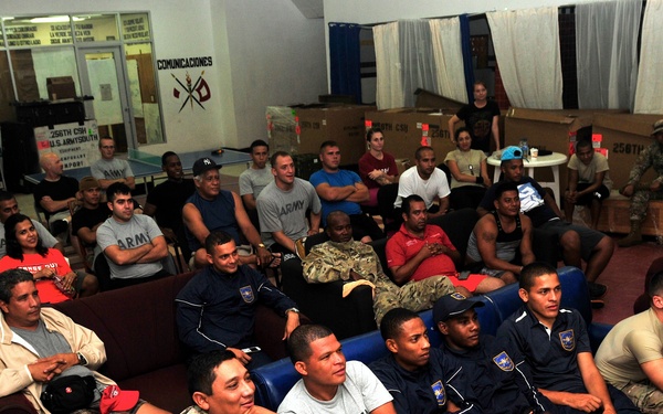 A large group of military personnel and civilians are gathered in what appears to be a common room or recreation area of a military facility. The group is seated on sofas and chairs, appearing to watch something off-camera, possibly a presentation or screen. The setting includes visible military signage in Spanish, including the word 'COMUNICACIONES,' suggesting a Latin American or Spanish-speaking military installation.
