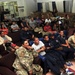 A large group of military personnel and civilians are gathered in what appears to be a common room or recreation area of a military facility. The group is seated on sofas and chairs, appearing to watch something off-camera, possibly a presentation or screen. The setting includes visible military signage in Spanish, including the word 'COMUNICACIONES,' suggesting a Latin American or Spanish-speaking military installation.
