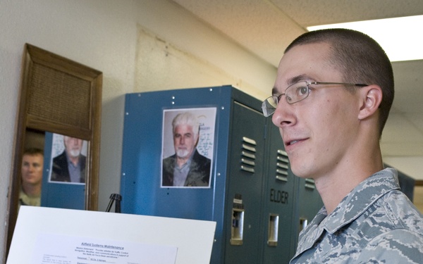 A U.S. Air Force airman in ABU (Airman Battle Uniform) is presenting a briefing board displaying information about 'Airfield Systems Maintenance' and 'Air Traffic Communication Systems'. The presentation appears to be taking place in a hallway or classroom setting with lockers visible in the background. Another military personnel member is partially visible in the background observing the presentation.