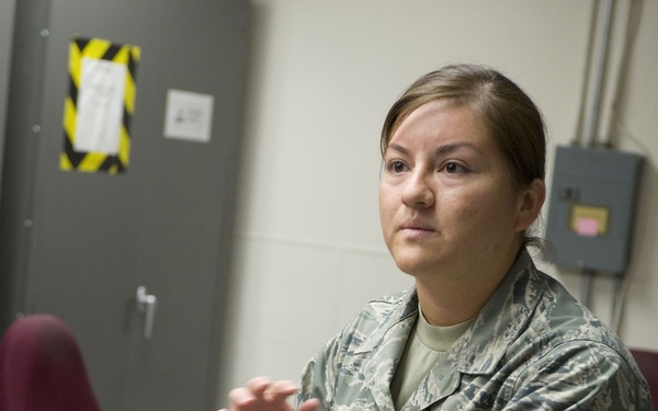 A U.S. Air Force female service member in ABU (Airman Battle Uniform) camouflage is standing at a table handling what appears to be a harness or safety equipment with red and black straps and metal D-rings. Her rank insignia on the sleeve indicates she is a Senior Airman (E-4), and her name tape partially reads 'DALE'. She appears to be explaining or demonstrating the equipment in an indoor facility.