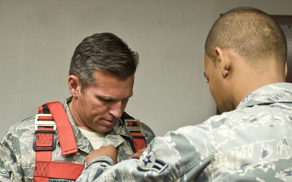 Two U.S. Air Force personnel in ABU (Airman Battle Uniform) camouflage are shown in what appears to be an indoor training or briefing room. One airman is assisting the other in fitting or adjusting a red safety harness over his uniform. The name tape 'CORNISH' (partially visible) is readable on the airman wearing the harness.