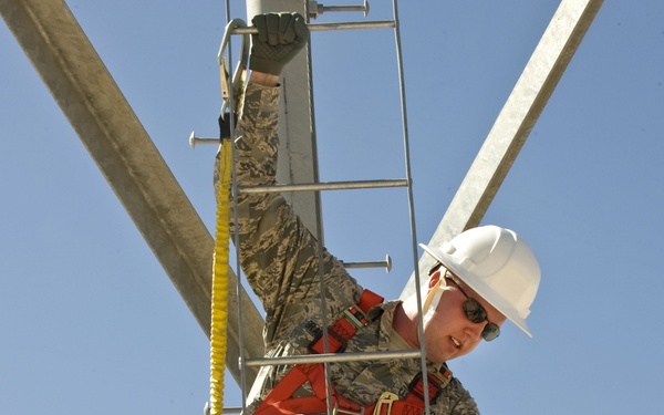 A military service member in US Air Force ABU (Airman Battle Uniform) camouflage is climbing a metal utility or communications tower. The individual is equipped with full fall-protection safety gear including a red harness, yellow lanyard, white hard hat, safety glasses, and gloves. The photo is taken from a low angle looking upward, emphasizing the height and the structural elements of the tower against a clear blue sky.