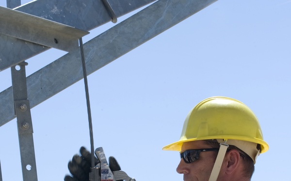 A military service member in US Air Force ABU (Airman Battle Uniform) camouflage is climbing or working on a metal structure at height. The individual is equipped with full fall protection safety gear including a yellow hard hat, safety harness, gloves, and a rope grab/fall arrest device attached to a vertical lifeline. The background shows an arid, desert-like landscape with hills and clear blue sky.