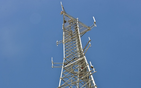 A ground-level upward-looking photograph of a tall lattice telecommunications tower against a clear blue sky. A single worker wearing a yellow hard hat and safety harness is visible climbing the tower structure at approximately one-third of the way up. The tower is equipped with multiple antennas, cable runs, and mounting hardware at various levels.