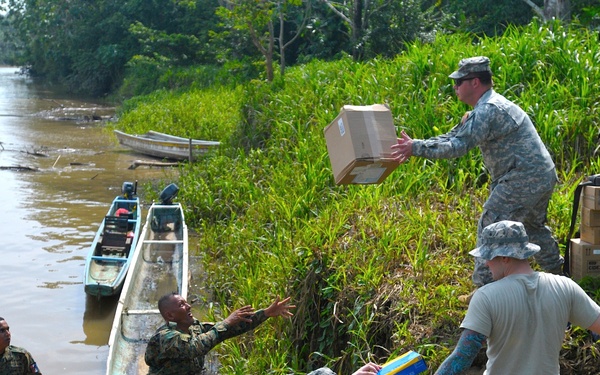 Military personnel in camouflage uniforms are conducting a supply unloading operation along a riverbank in what appears to be a tropical jungle environment. Soldiers from at least two different military forces are forming a human chain to pass cardboard boxes and supplies from boats moored at the muddy riverbank up to a staging area on higher ground. The operation appears to be a humanitarian aid or logistics resupply mission given the quantity of boxed goods visible.