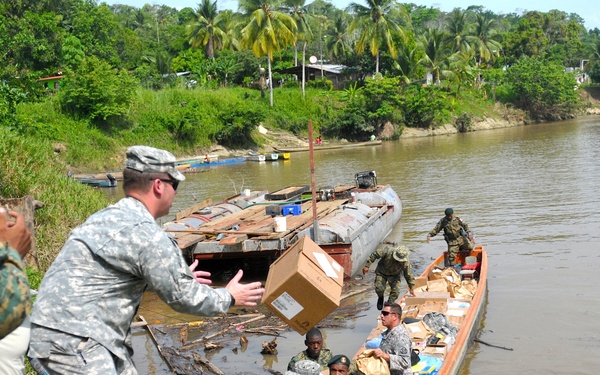 Military personnel from what appear to be multiple nations are conducting a humanitarian aid or logistics operation along a tropical river. Soldiers are unloading and passing cardboard boxes from large wooden dugout canoes to the riverbank. The scene suggests a coordinated supply distribution effort in a remote, jungle-bordered waterway.