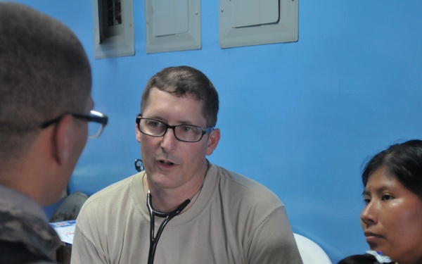 A medical professional wearing a stethoscope and glasses appears to be conducting a consultation with a woman and young child in a room with blue walls. A person in military camouflage uniform is present, suggesting this may be a military humanitarian or medical assistance mission. The setting appears to be a makeshift or temporary medical clinic.