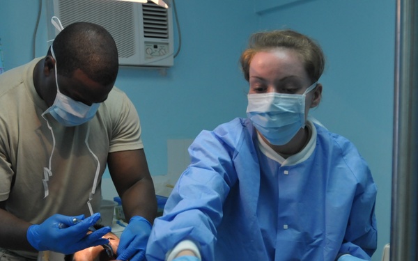 Two medical/dental professionals wearing surgical masks, gloves, and protective gowns are performing a dental procedure on a patient reclined in a dental chair. One provider in military-style clothing is working directly in the patient's mouth while the other in a blue surgical gown is reaching for instruments on a tray. The setting appears to be a basic dental clinic with blue walls, a window AC unit, and standard dental equipment.