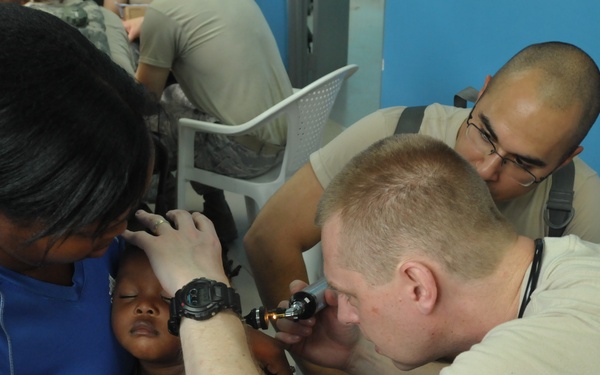 A medical examination is being conducted in what appears to be a field clinic or humanitarian mission setting. A military or medical personnel member is using an otoscope to examine a young child's ear while a woman, likely the child's mother, holds the child steady. A second uniformed individual observes the procedure from behind.