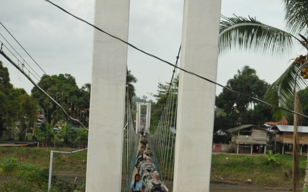 A group of military personnel in camouflage uniforms are crossing a suspension footbridge over a river in what appears to be a tropical, rural setting. At least one civilian, appearing to be a young woman in a blue shirt and dark skirt, is also on the bridge among the soldiers. The surrounding environment features palm trees, dense vegetation, wooden structures, and overcast skies consistent with a Central or South American locale.