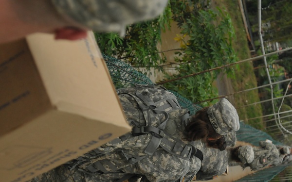 The image shows U.S. Army soldiers in ACU (Army Combat Uniform) camouflage handling cardboard boxes, likely during a humanitarian aid or supply distribution operation. The photo is taken at an unusual tilted angle, showing soldiers passing or stacking boxes along what appears to be a wall or barrier with green netting. The background shows a tropical or subtropical environment with lush green vegetation.