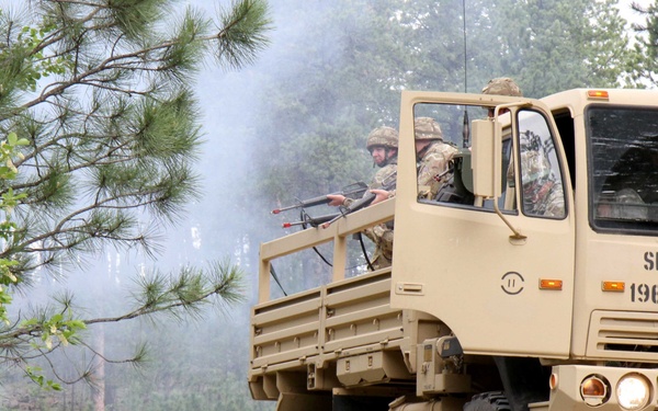 A military training exercise is underway in a forested area, featuring a tan LMTV (Light Medium Tactical Vehicle) marked 'SDNG 196MEB' belonging to the South Dakota National Guard. Soldiers in combat gear are positioned in and around the vehicle, with one individual in civilian-style white clothing throwing an object toward the convoy, simulating an ambush or IED scenario. A Humvee is visible in the background amid smoke.