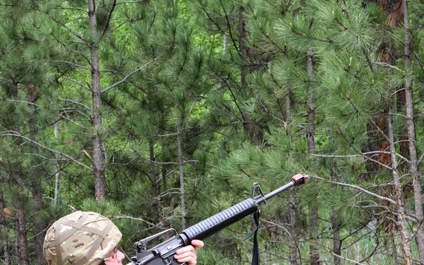 A soldier in MultiCam/MTP camouflage uniform is crouching in a kneeling position and aiming an M16-style rifle upward at an angle in a forested environment. The individual is wearing full combat kit including helmet, load-bearing equipment with pouches, and black combat boots. The setting appears to be a training exercise based on the visible orange muzzle safety device on the rifle barrel.