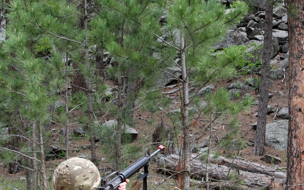 A soldier in MultiCam/MTP camouflage uniform is crouched in a kneeling position aiming a rifle upward in a forested, rocky terrain. The individual is equipped with full combat gear including a helmet, load-bearing vest with pouches, and black boots. The setting appears to be a training exercise or field operation in a pine forest environment.