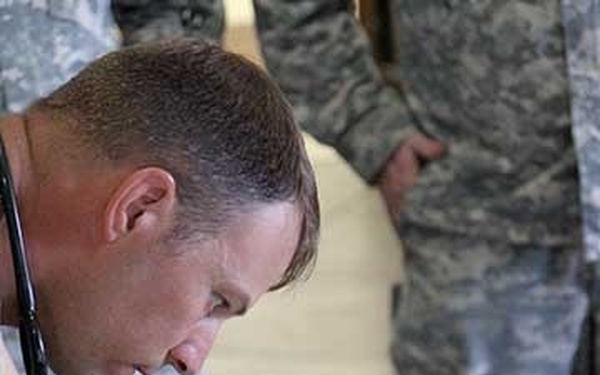 A man wearing a stethoscope around his neck is leaning over a table, writing on a clipboard or form. He appears to be a medical professional working in a military setting, as individuals in camouflage uniforms are visible in the background. A disposable coffee cup is on the table beside him.