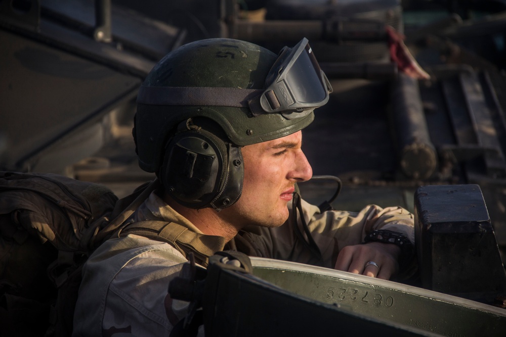 A military service member is positioned inside or atop a military vehicle, wearing combat gear and looking to the right. The soldier is equipped with a tactical helmet, protective goggles, hearing protection headset, desert camouflage uniform, and a tactical vest. A serial number '533430' is visible on the vehicle's metal surface.