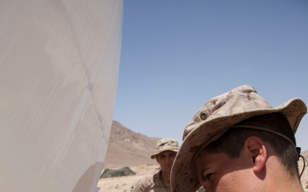 Two military personnel in desert camouflage uniforms (MARPAT desert pattern) are working in an arid, desert environment. The foreground soldier, wearing a boonie hat and displaying sergeant chevrons on his sleeve, is operating a handheld radio device near what appears to be a white fabric structure or antenna reflector. A second soldier observes from the background in a sandy, mountainous terrain.