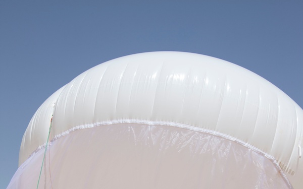 A group of military personnel in desert camouflage uniforms are preparing and inflating a large white tethered aerostat or surveillance balloon in an arid desert environment. Several soldiers are working together to manage the balloon's envelope and attach payload equipment underneath. The setting appears to be a military training or operational exercise in a desert location.